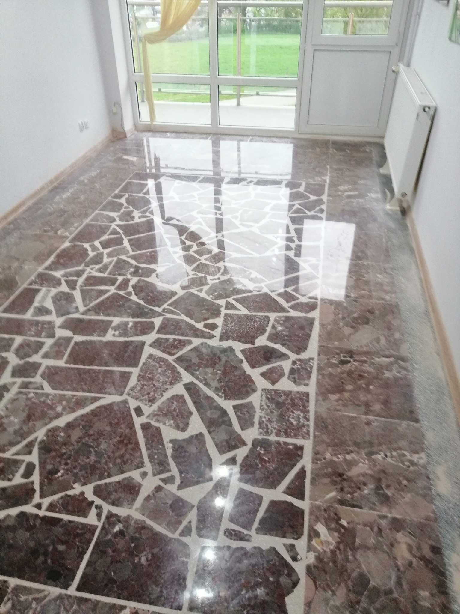 Błyszcząca posadzka z nieregularnych, brązowo-bordowych płytek z białymi fugami, odbijająca światło z okna balkonowego w pustym pomieszczeniu z białymi ścianami i grzejnikiem.