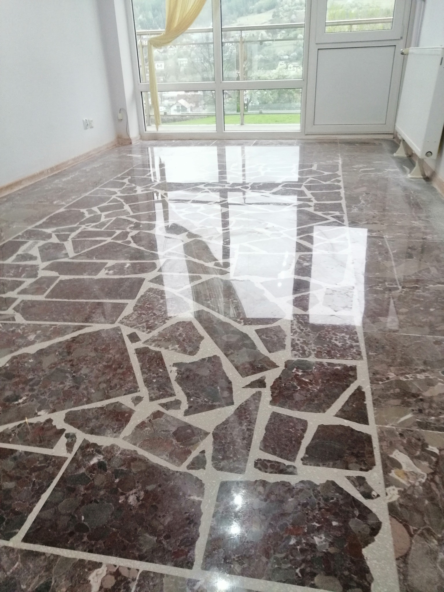 Błyszcząca podłoga z brązowych, nieregularnych płyt kamiennych z jasnymi fugami, odbijająca światło z okna balkonowego.