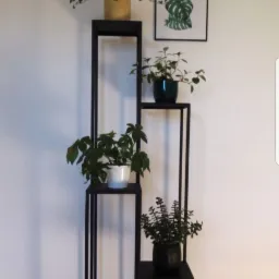 Metalowa konstrukcja schodkowa w kolorze czarnym, służąca jako kwietnik, z czterema półkami na których ustawione są doniczki z różnymi roślinami, na ścianie za konstrukcją wisi obrazek z motywem...