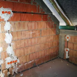 Montaż przewodów elektrycznych w bruzdach w ścianie z czerwonej cegły, gniazdka elektryczne zabezpieczone gipsem, widoczne przewody prowadzone wzdłuż ściany po podłodze.