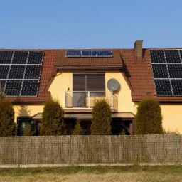 Żółty dom jednorodzinny z czerwoną dachówką i panelami fotowoltaicznymi na dachu, widoczny balkon z satelitą i mały ptak na barierce, otoczony tujami i bambusowym płotem.