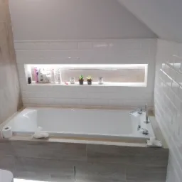 Wanna obudowana płytkami imitującymi drewno, białe płytki typu metro na ścianach, wnęka z oświetleniem i kosmetykami, minimalistyczna aranżacja łazienki na poddaszu.