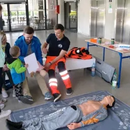 Instruktor w pomarańczowym uniformie demonstruje techniki ratownicze na manekinie przed grupą osób, w tym dzieckiem trzymającym kartkę.