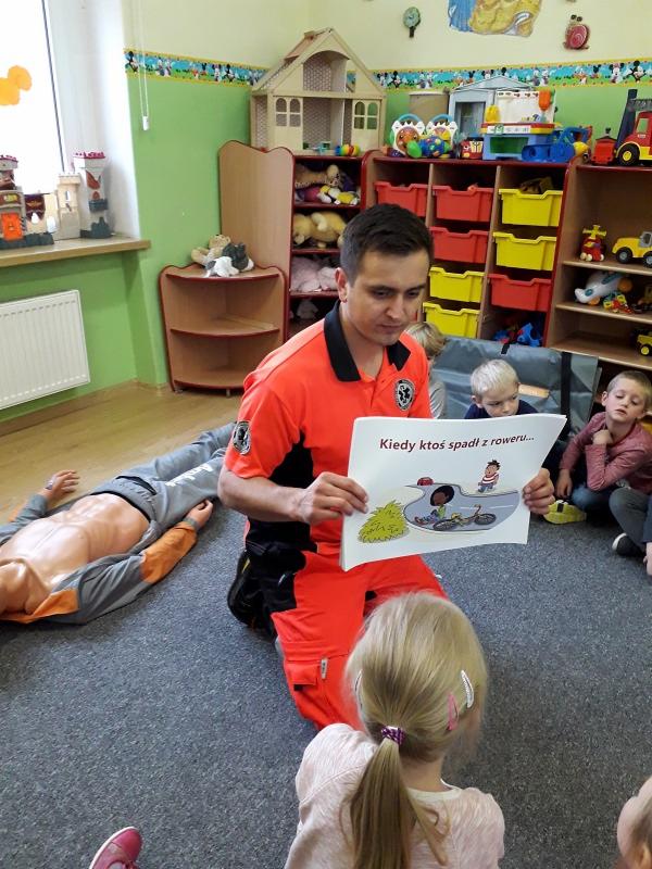 Instruktor w pomarańczowym uniformie klęczy, prezentując ilustrację o bezpieczeństwie rowerowym dzieciom podczas zajęć w sali przedszkolnej, obok leży manekin do ćwiczeń resuscytacji.