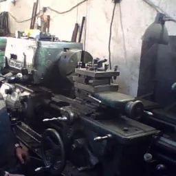 Operator z wąsami w czapce, pozuje obok tokarki w warsztacie oświetlonym lampą na giętkim ramieniu.