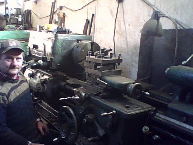 Operator z wąsami w czapce, pozuje obok tokarki w warsztacie oświetlonym lampą na giętkim ramieniu.