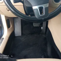 Wnętrze samochodu z czarnym dywanikiem pod pedałami, widok z góry na kierownicę i beżowe elementy deski rozdzielczej.