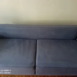 Szara, dwuosobowa kanapa po czyszczeniu tapicerki, widoczne delikatne różnice w odcieniu na siedzisku, jasne tło ściany, parkiet na podłodze.