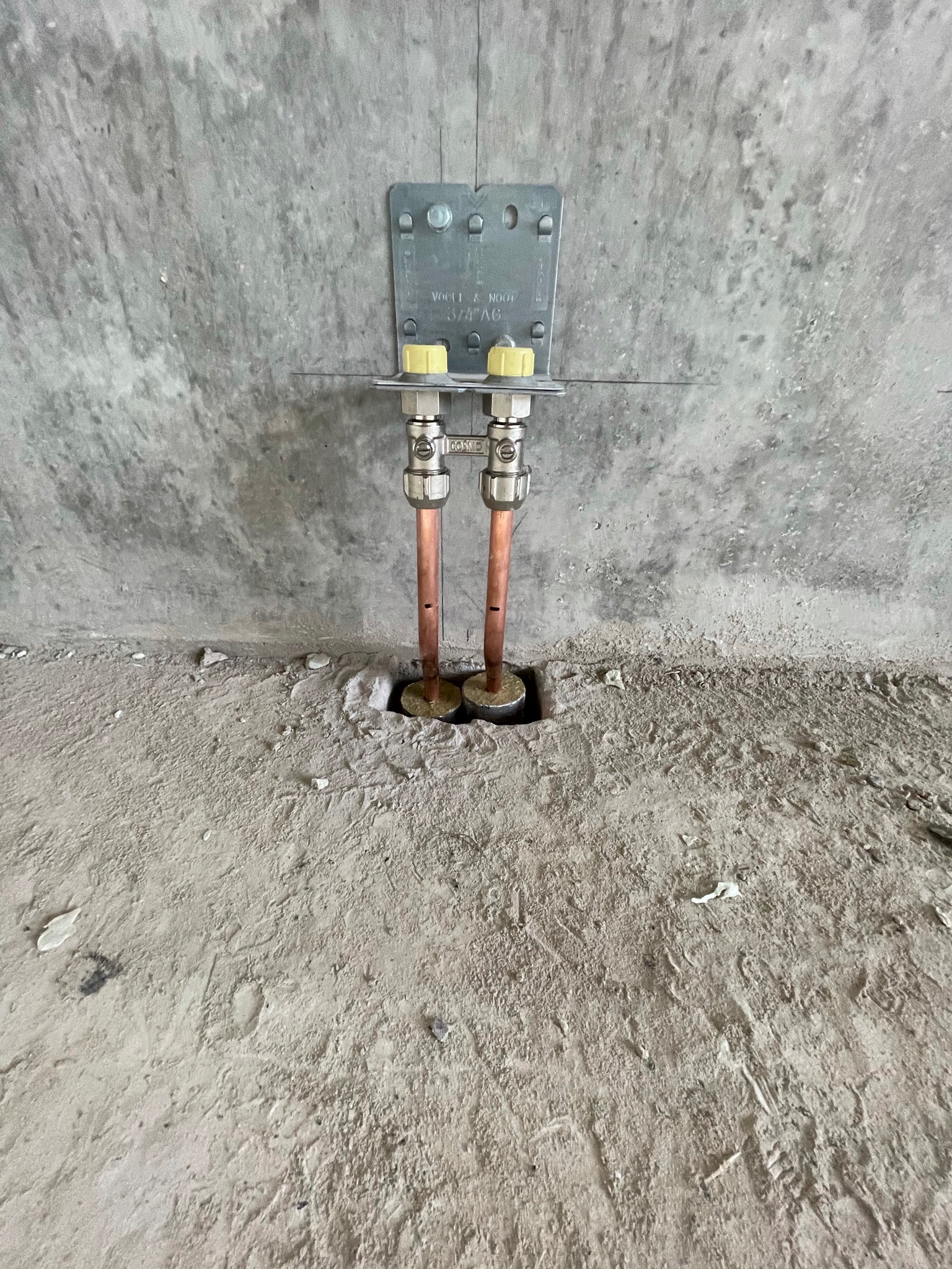 Miedziane rury instalacji wodnej wystające ze ściany i podłogi w surowym stanie, zamocowane do metalowej płyty z żółtymi zaworami.