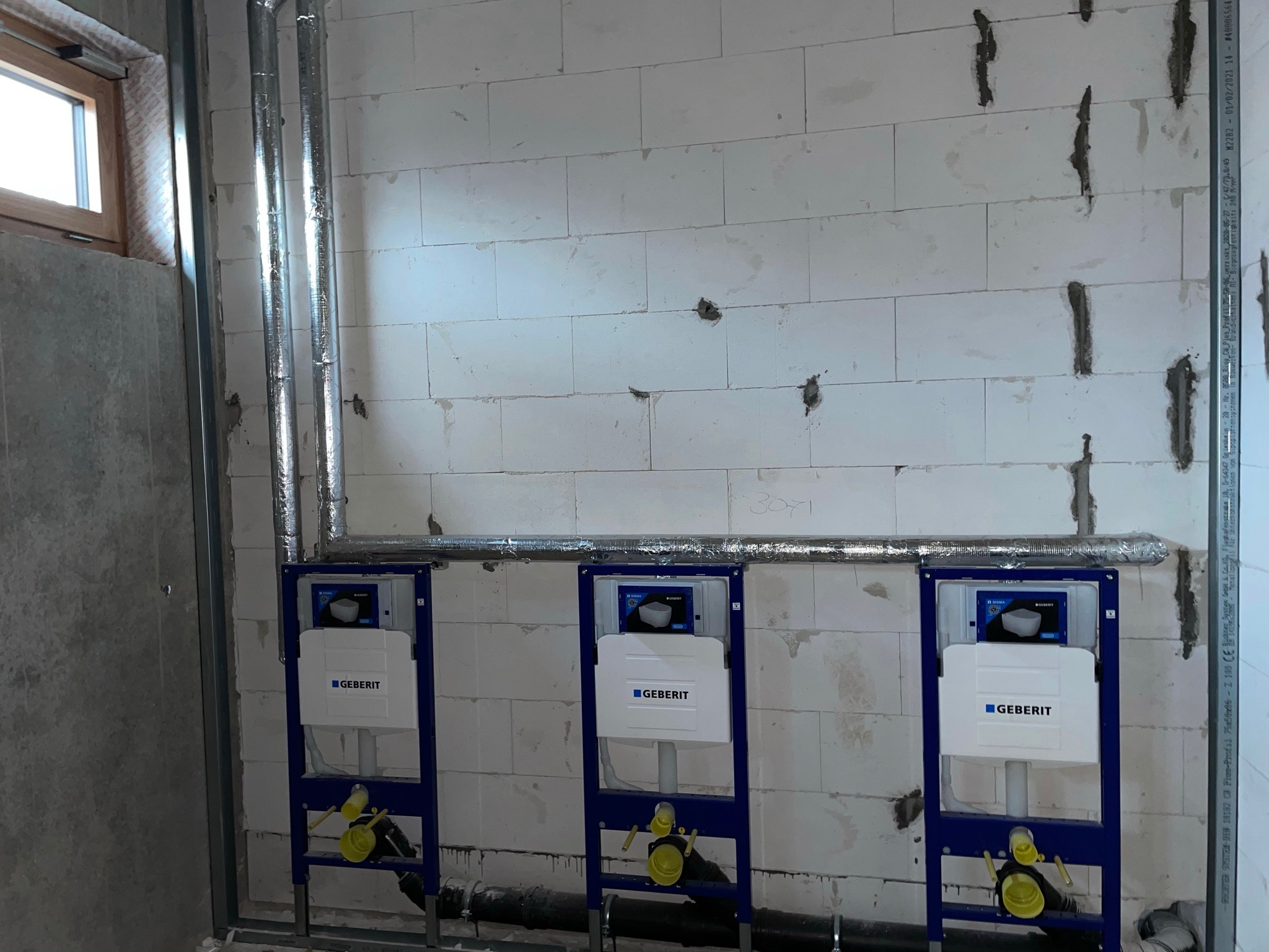 Trzy stelaże podtynkowe Geberit zamontowane przy ścianie z betonu komórkowego, widoczne rury instalacyjne w izolacji, surowy stan wykończenia.