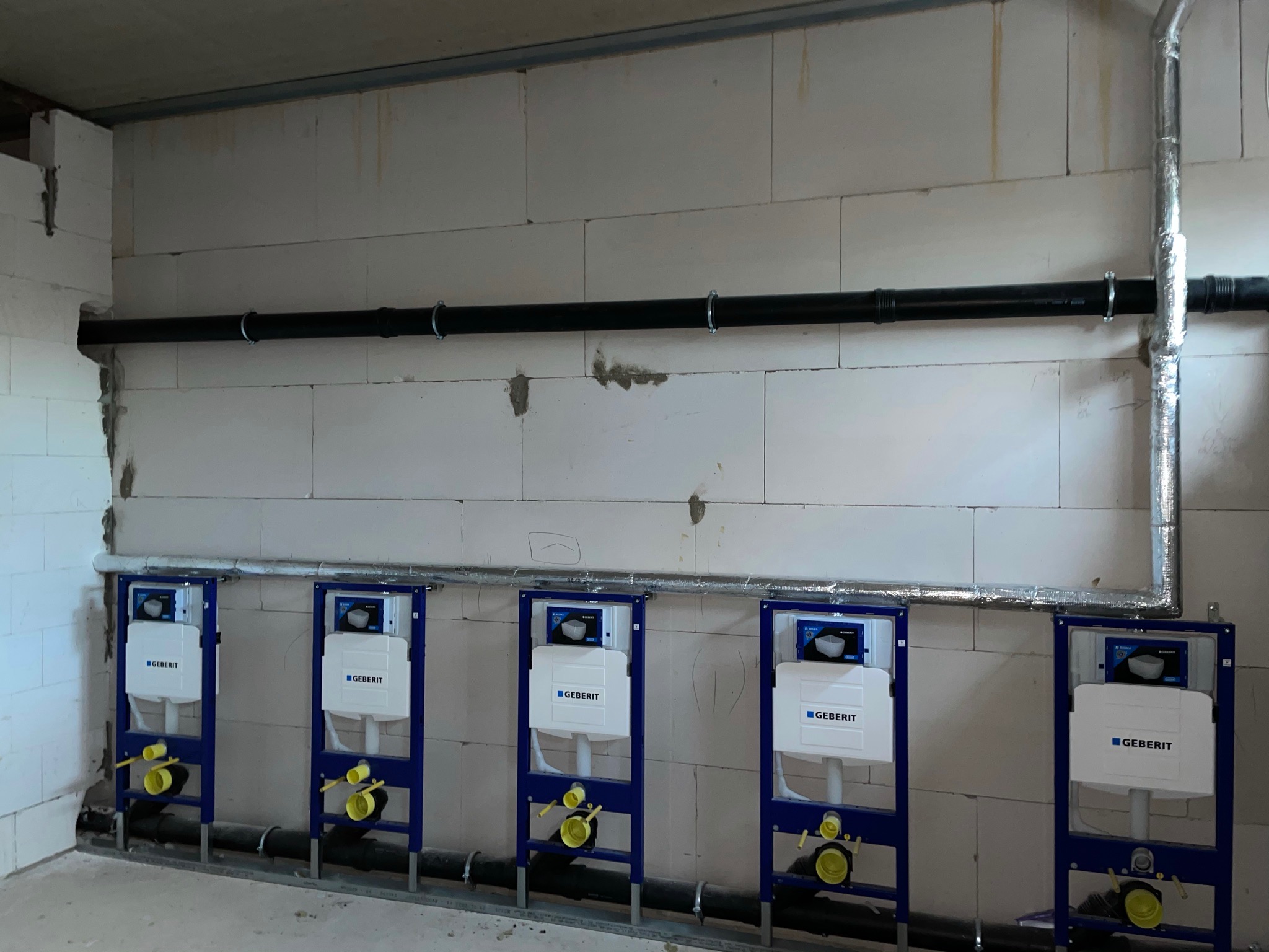 Rząd pięciu stelaży podtynkowych Geberit zamontowanych na ścianie z betonu komórkowego, z widocznymi rurami odpływowymi i dopływowymi, instalacja w trakcie realizacji.