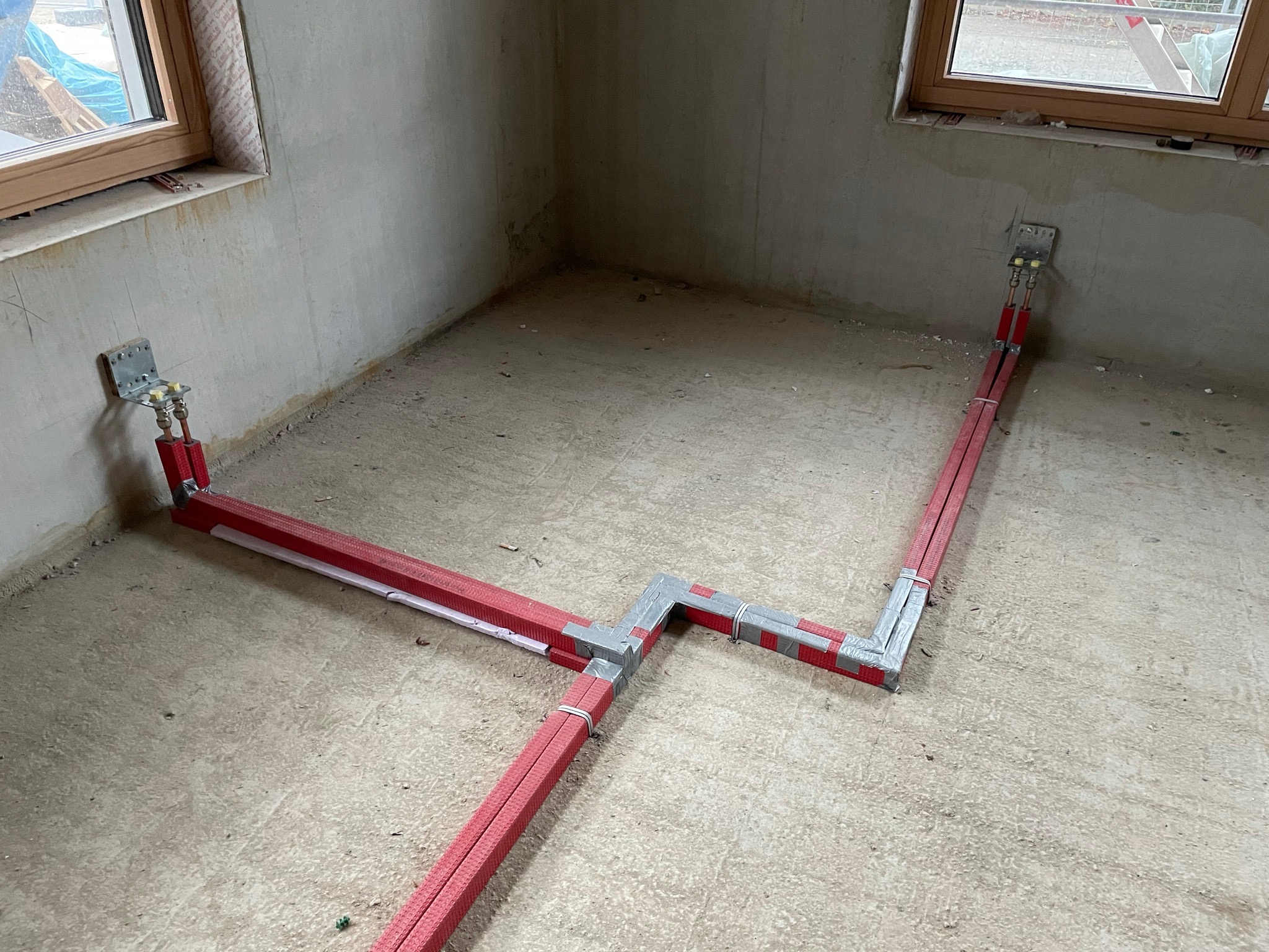 Nowa instalacja grzewcza podłogowa w surowym stanie, rury w czerwonej izolacji ułożone na posadzce, mocowane do ściany metalowymi uchwytami.