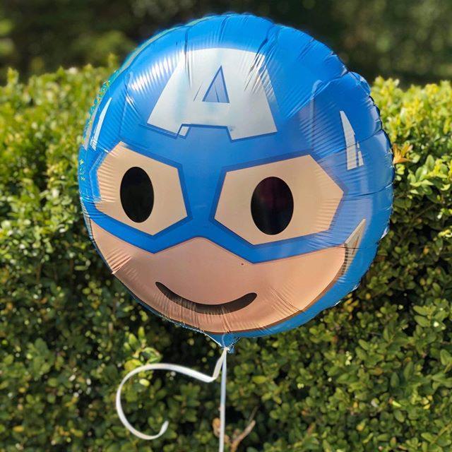 Niebieski balon foliowy z nadrukiem twarzy superbohatera z dużym A na czole, na tle zielonych krzewów.