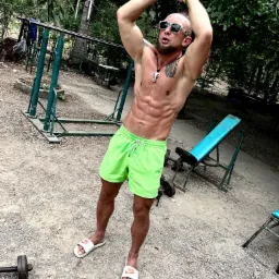 Mężczyzna w neonowych szortach i okularach przeciwsłonecznych, prezentujący wyrzeźbione ciało na tle plenerowej siłowni z zardzewiałymi sprzętami i drzewami, w trakcie rozciągania rąk nad głową.
