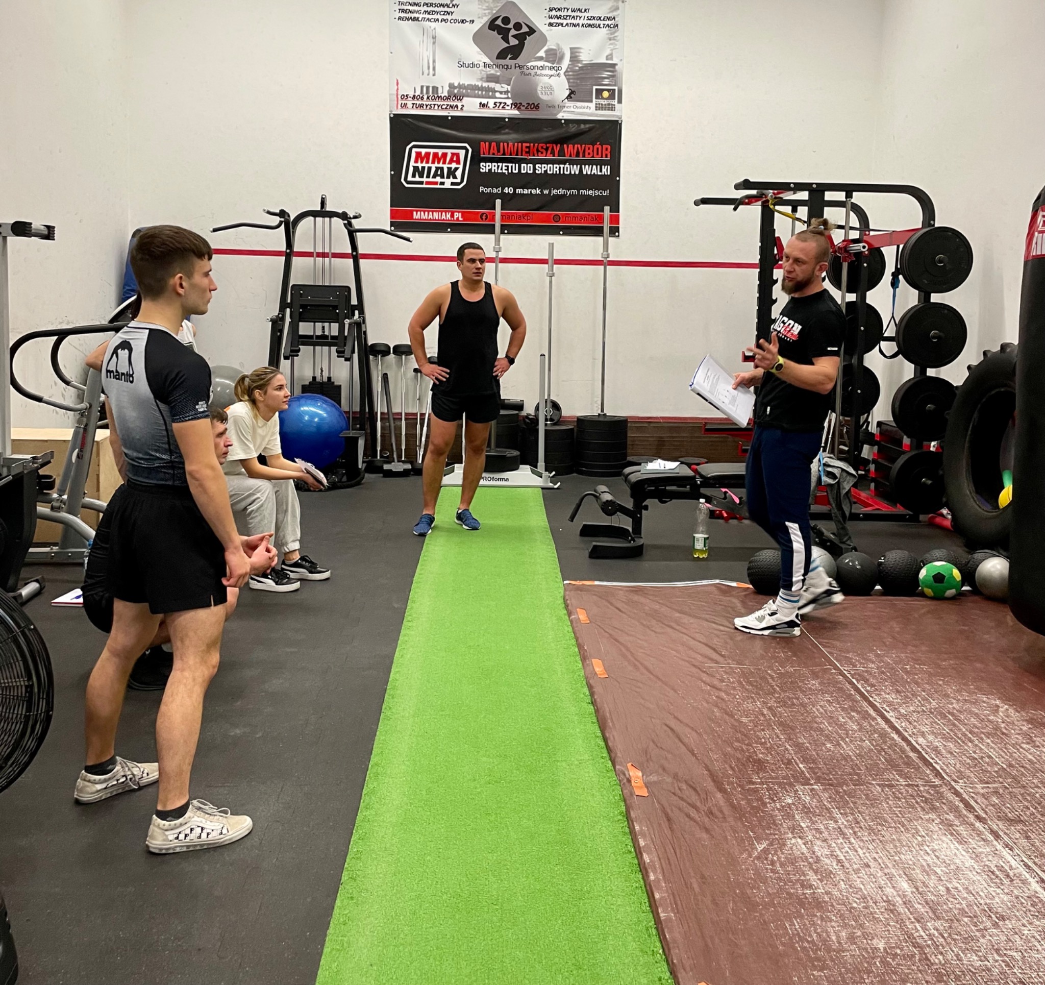 Grupa osób w siłowni podczas instruktażu, trener z brodą i kartką w ręku objaśnia ćwiczenia, widoczny sprzęt do ćwiczeń siłowych i worki treningowe.