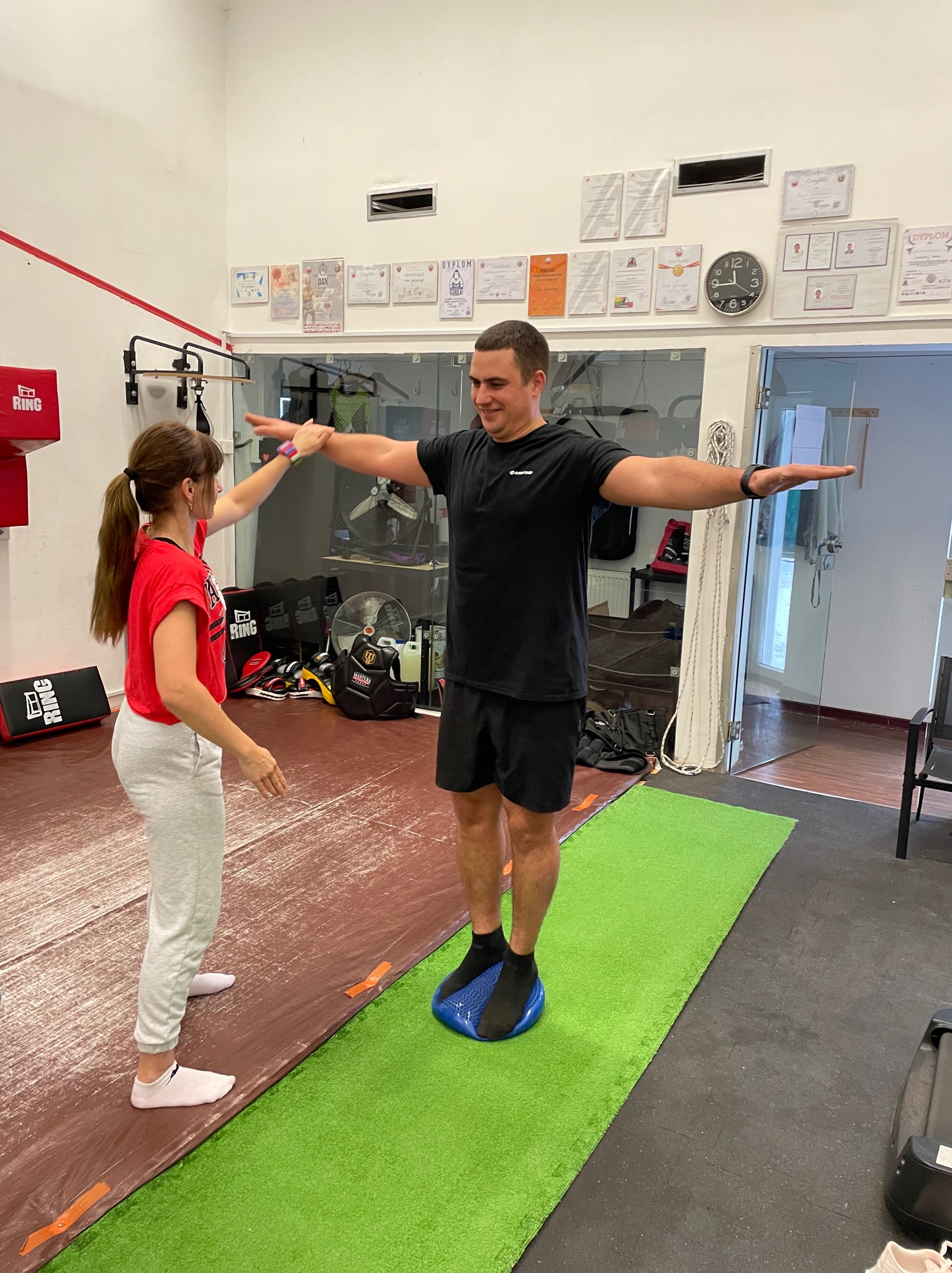 Mężczyzna w czarnej koszulce i spodenkach stoi na niebieskiej poduszce sensorycznej, z rozłożonymi ramionami, podczas gdy kobieta w czerwonej koszulce sprawdza jego postawę w jasnym pomieszczeniu...