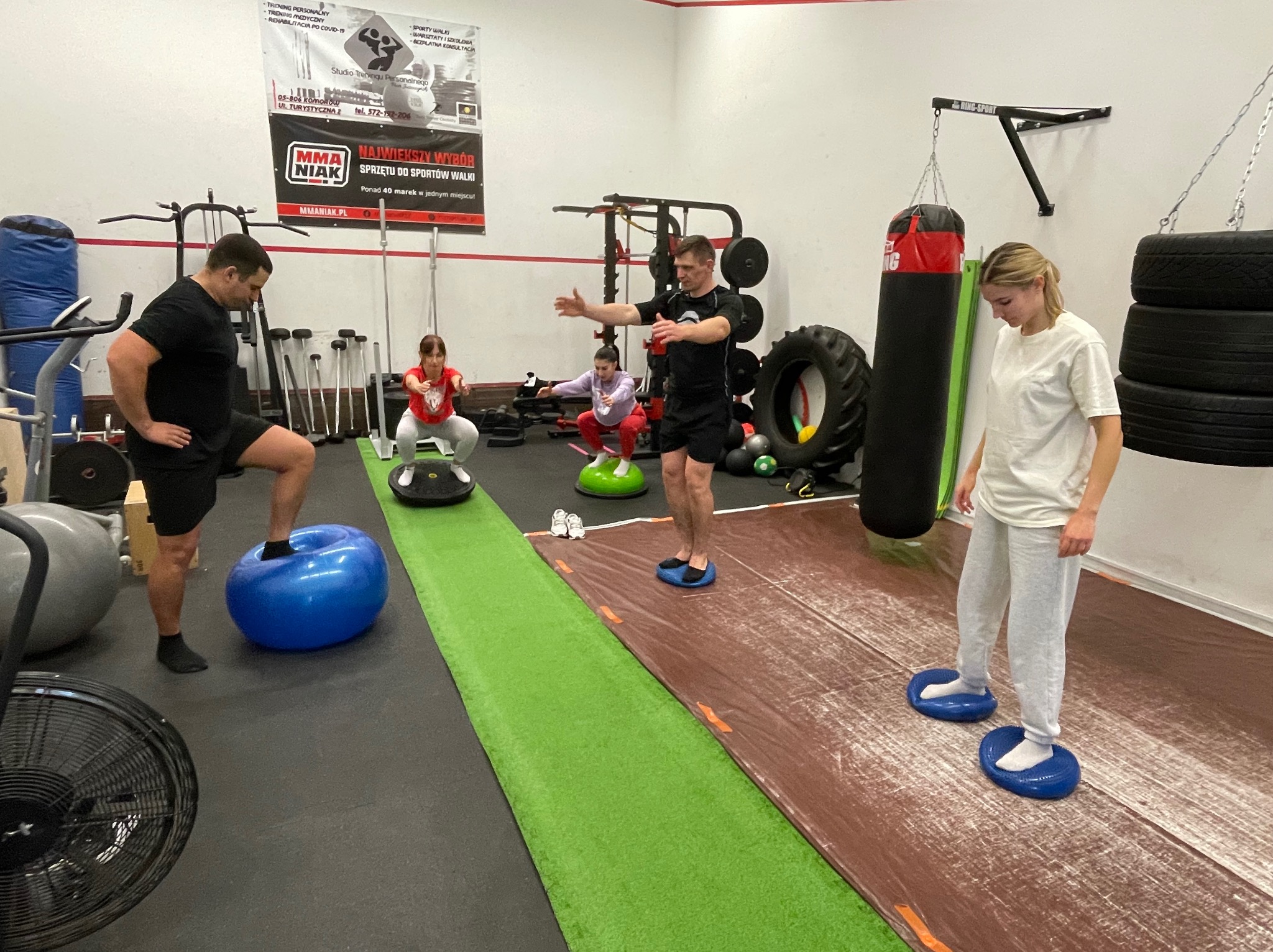 Grupa osób ćwiczy równowagę na różnych przyrządach w przestronnej sali treningowej z plakatem MMA Niak na ścianie.
