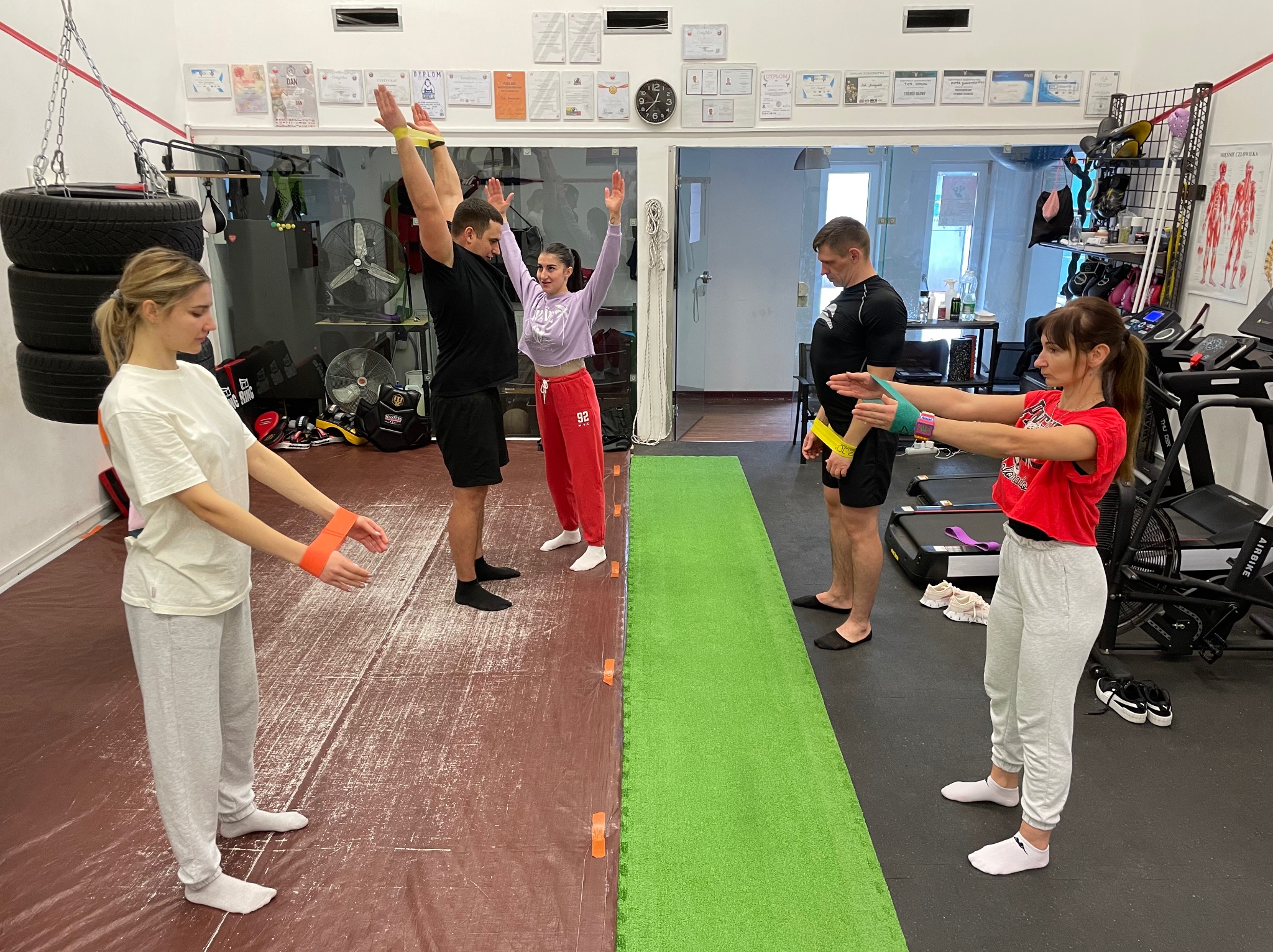 Grupa osób ćwiczy z gumami oporowymi w przestronnej sali gimnastycznej z lustrami, dyplomami na ścianach i sprzętem do ćwiczeń w tle.
