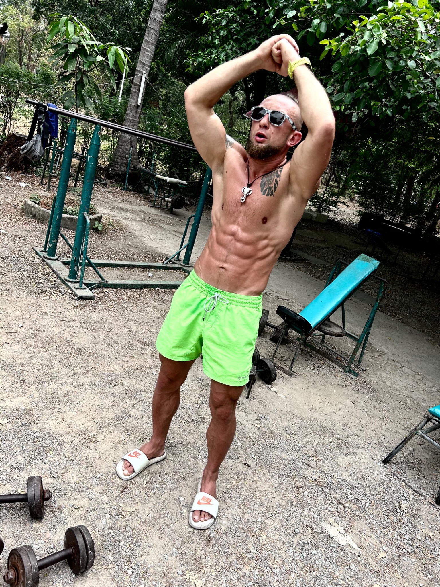 Mężczyzna w neonowych szortach i okularach przeciwsłonecznych, prezentujący wyrzeźbione ciało na tle plenerowej siłowni z zardzewiałymi sprzętami i drzewami, w trakcie rozciągania rąk nad głową.