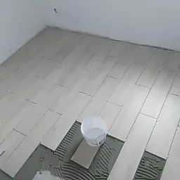 Układanie jasnych, podłużnych płytek podłogowych z krzyżykami dystansowymi w pomieszczeniu; widoczny kubeł z wodą i zaprawa klejowa.