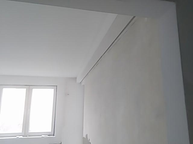 Białe ściany w trakcie malowania, widoczne niedociągnięcia i nierówności, okno z jasnym światłem dziennym w tle, surowy stan wykończenia wnętrza.