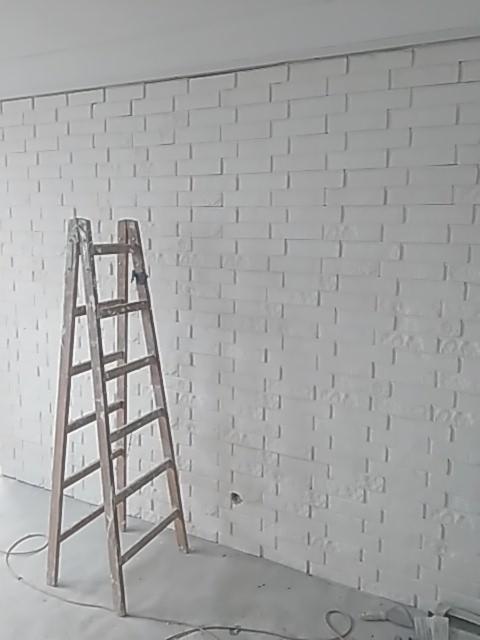 Świeżo pomalowana na biało ściana z cegieł dekoracyjnych, drewniana drabina oparta o ścianę, widoczne ślady farby na podłodze i kablu zasilającym.
