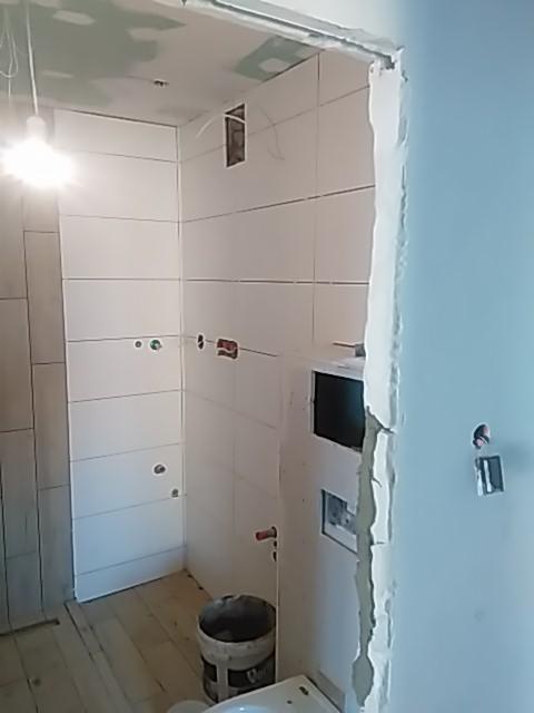 Wnętrze łazienki w trakcie wykańczania: białe płytki na ścianach, widoczne otwory na instalacje, surowe drewniane podłogi, plastikowe wiadro i niedokończone obramowanie drzwi.