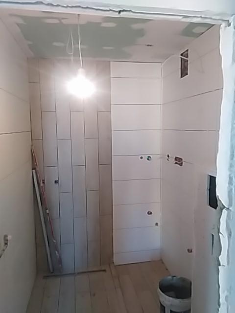 Wnętrze łazienki w trakcie remontu: białe, poziome płytki ułożone na ścianie, surowy sufit z widocznymi łatami gipsowymi, drewniana podłoga, drabina oparta o ścianę, widoczne otwory na instalacje...