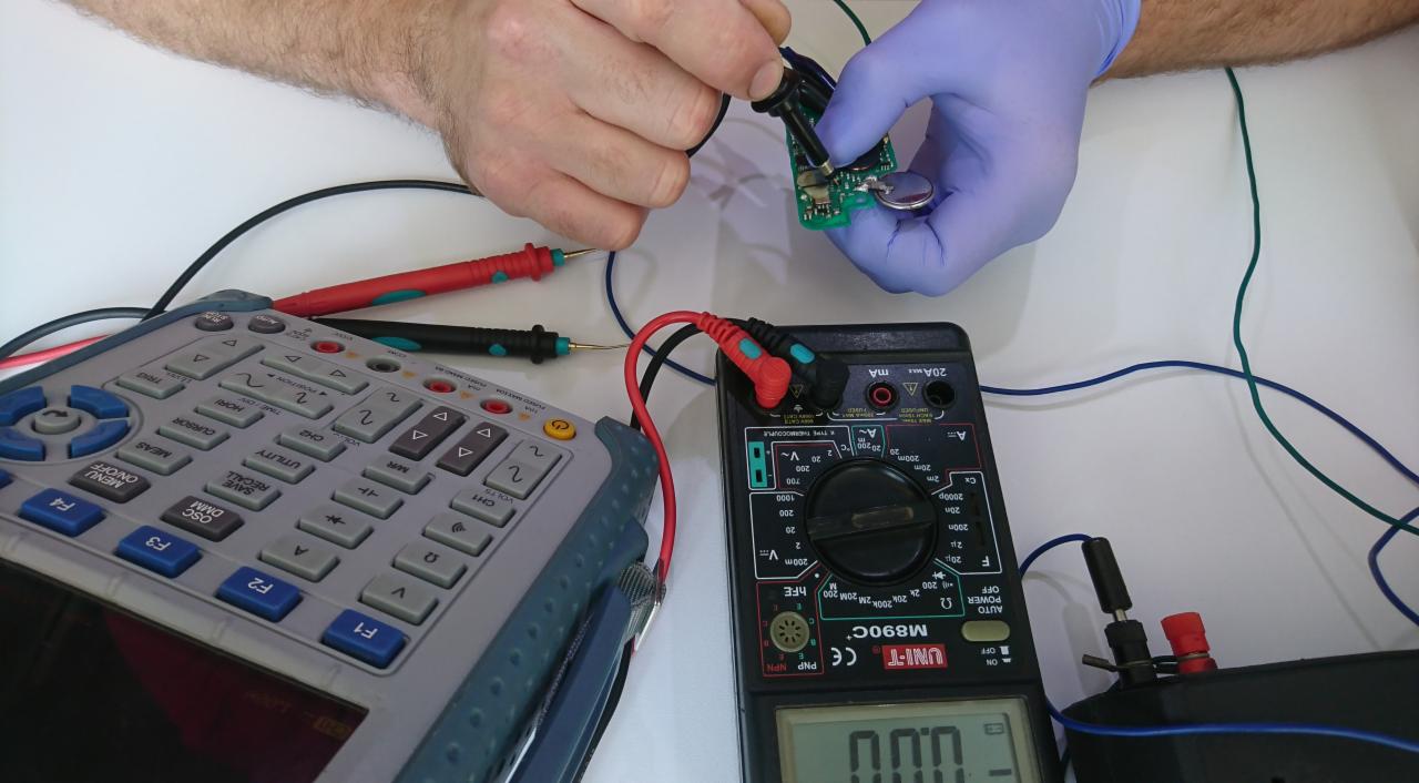 Diagnostyka elektroniczna: Osoba w rękawiczkach ochronnych mierzy napięcie na płytce drukowanej z baterią pastylkową za pomocą sondy, obok widoczny oscyloskop i multimetr.