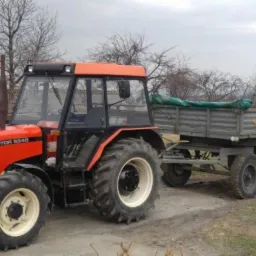 Czerwony traktor Zetor 5340 z przyczepą na tle pola i drzew bez liści, pochmurne niebo.