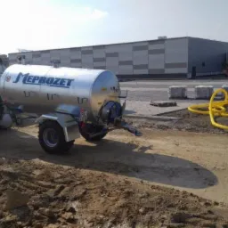 Czerwony traktor z srebrną cysterną firmy Meprozet na placu budowy, w tle budynek magazynowy z napisem STO i żółte rury.