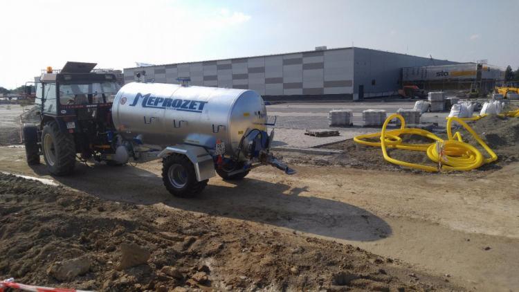 Czerwony traktor z srebrną cysterną firmy Meprozet na placu budowy, w tle budynek magazynowy z napisem STO i żółte rury.