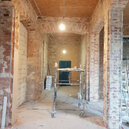 Wnętrze budynku w trakcie prac remontowych, widoczne ceglane kolumny z elementami wzmacniającymi, sufit z płyt OSB, instalacje elektryczne pod sufitem, rusztowanie na kółkach i worki z zaprawą w tle.