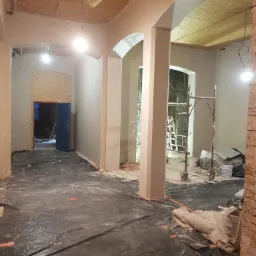 Wnętrze budynku w trakcie prac wykończeniowych, widoczne świeżo położone gładzie na ścianach i suficie z płyt OSB, podłoga zabezpieczona folią, pomieszczenie z łukowymi przejściami.