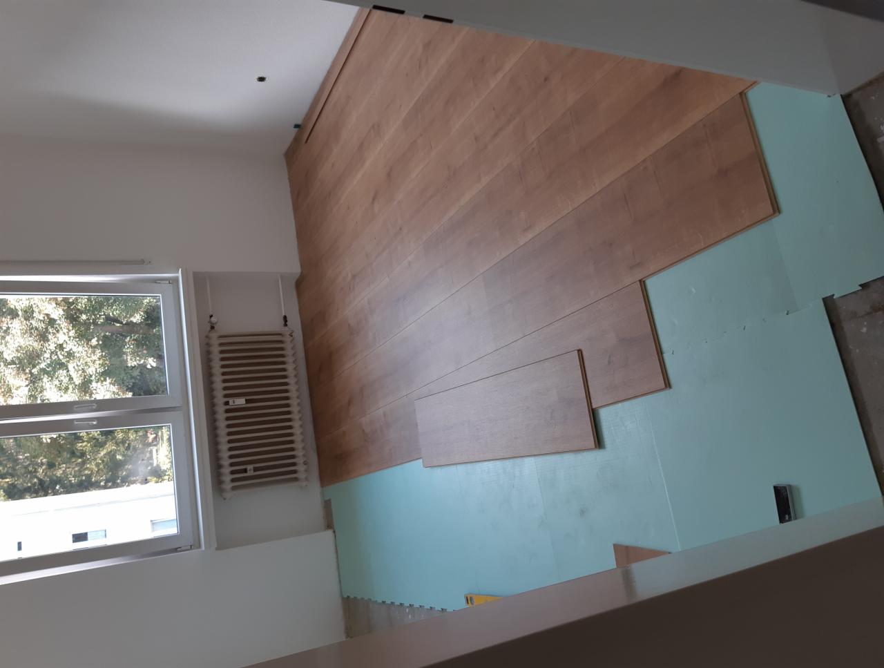 Układanie jasnych paneli podłogowych na zielonej macie izolacyjnej, widok z góry na fragment podłogi, grzejnik przy ścianie, okno w tle.