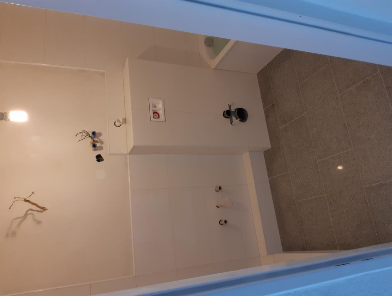 Łazienka w trakcie remontu: białe płytki ścienne, szare płytki podłogowe, wystające kable i rury przygotowane do podłączenia armatury, widoczne otwory na baterie i przycisk spłuczki.