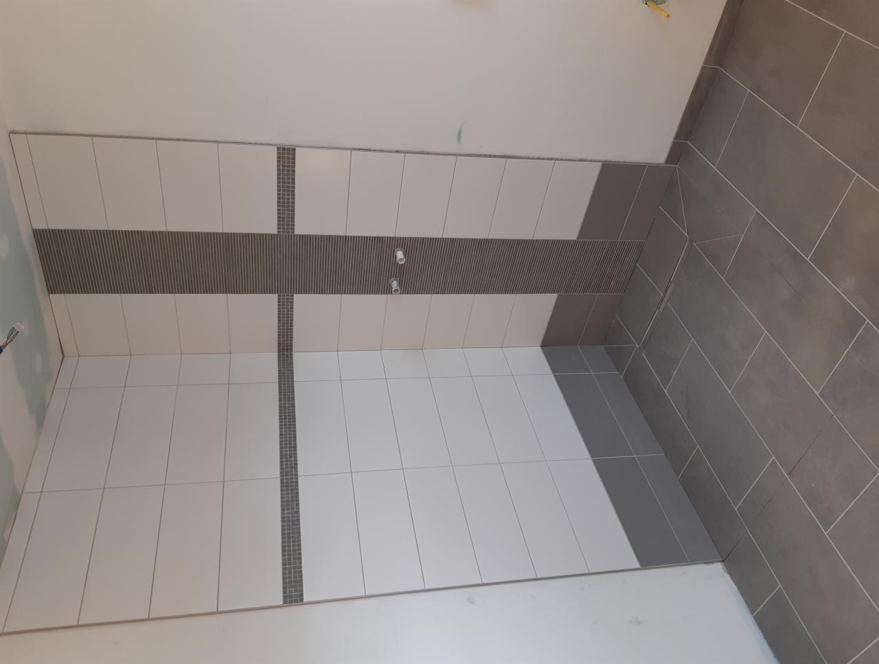 Świeżo ułożone, biało-szare płytki w łazience z dekoracyjnym pasem mozaiki i szarą podłogą z płytek imitujących kamień, widoczne dwa punkty instalacji wodnej.