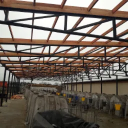 Konstrukcja stalowa z drewnianymi elementami pod zadaszenie nad betonową wylewką z licznymi budami dla zwierząt, w tle budynek gospodarczy i sprzęt budowlany.