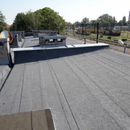 Szary, płaski dach pokryty papą z widocznymi kominami wentylacyjnymi i anteną satelitarną, w tle torowisko kolejowe i zieleń.