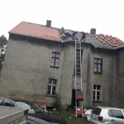 Ekipa dekarzy na dachu dwupiętrowego budynku z szarą elewacją, układająca czerwoną dachówkę; drabina oparta o ścianę, samochody zaparkowane przed budynkiem.