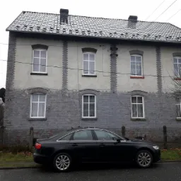 Dwupiętrowy dom z szarej cegły z ciemnym dachem i czarnym samochodem zaparkowanym przed nim w pochmurny dzień.