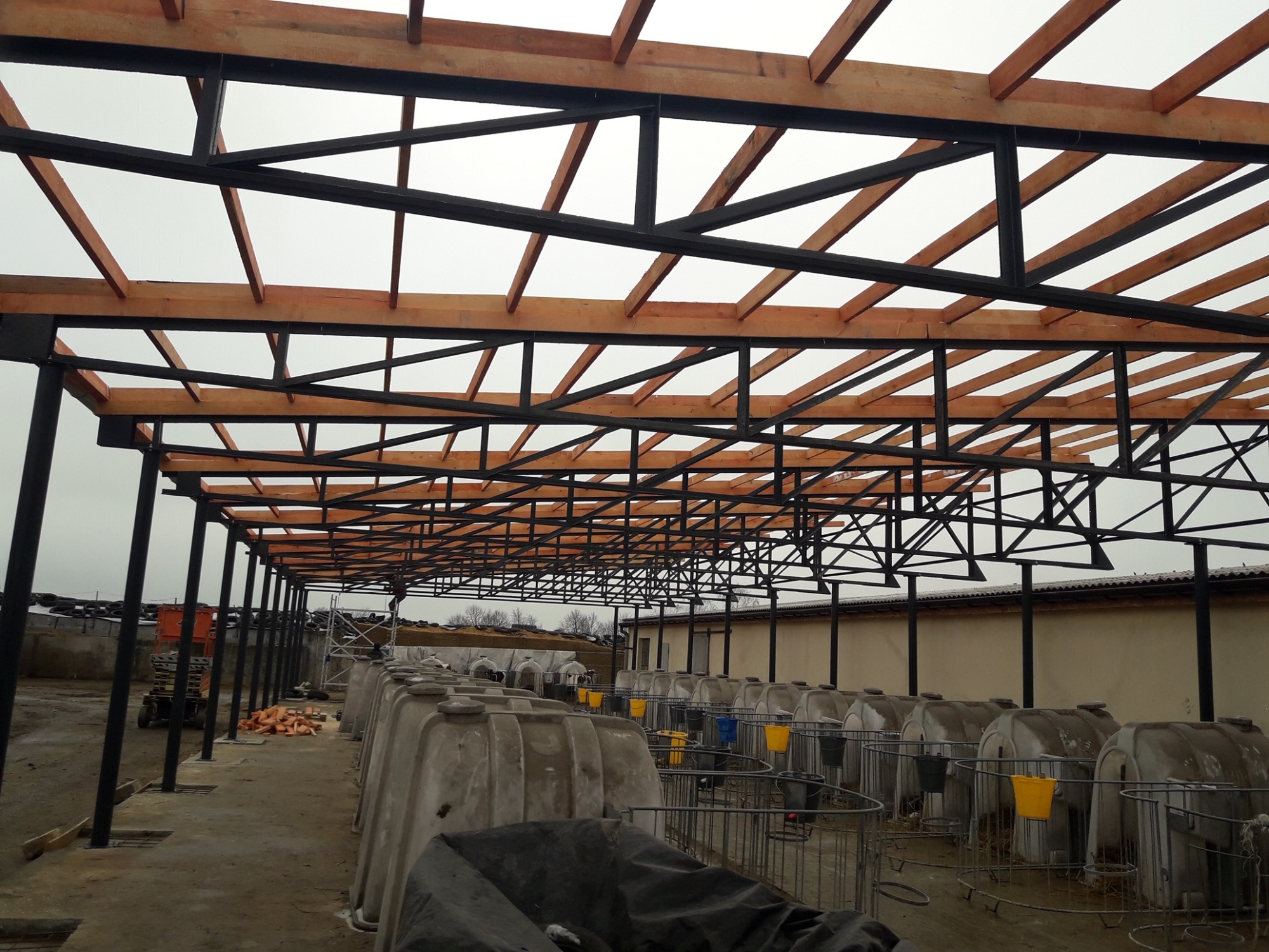 Konstrukcja stalowa z drewnianymi elementami pod zadaszenie nad betonową wylewką z licznymi budami dla zwierząt, w tle budynek gospodarczy i sprzęt budowlany.