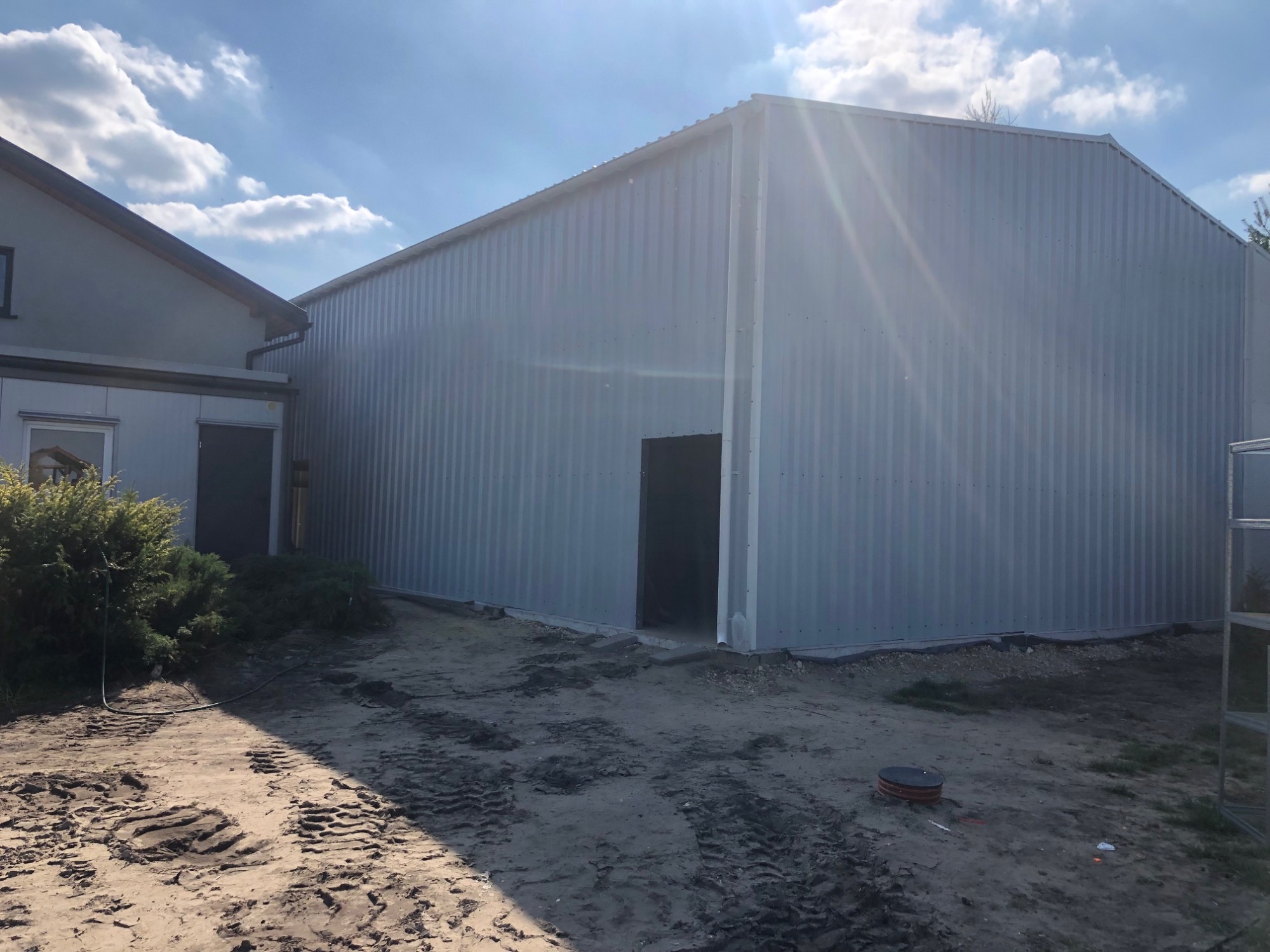 Nowo wybudowana hala z blachy trapezowej w słoneczny dzień, widok na boczną ścianę i wejście, obok budynek mieszkalny.