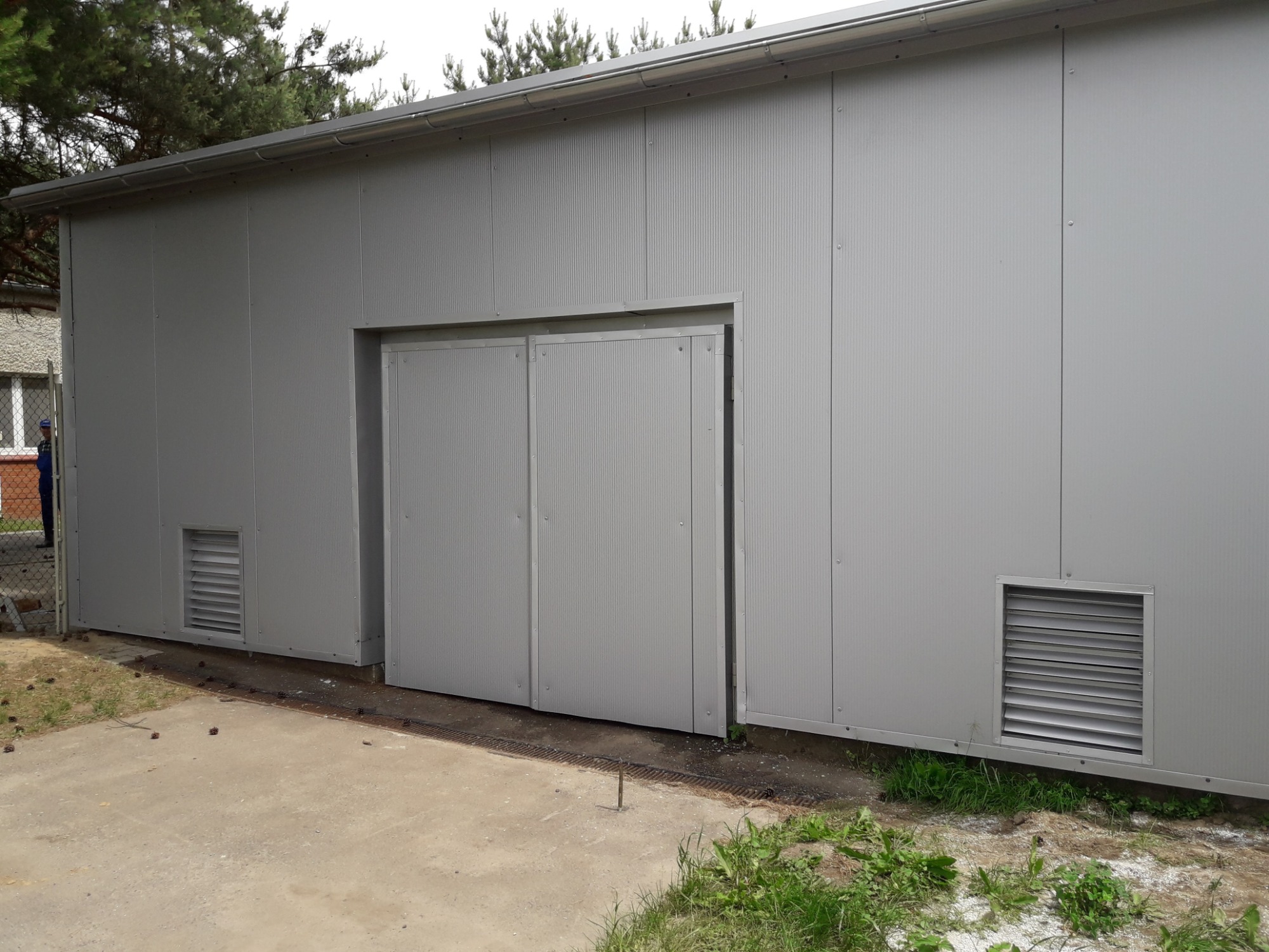 Szara hala magazynowa z panelowych płyt warstwowych, z dwuskrzydłowymi drzwiami i kratkami wentylacyjnymi, widok z zewnątrz na betonową posadzkę i fragment budynku mieszkalnego w tle.
