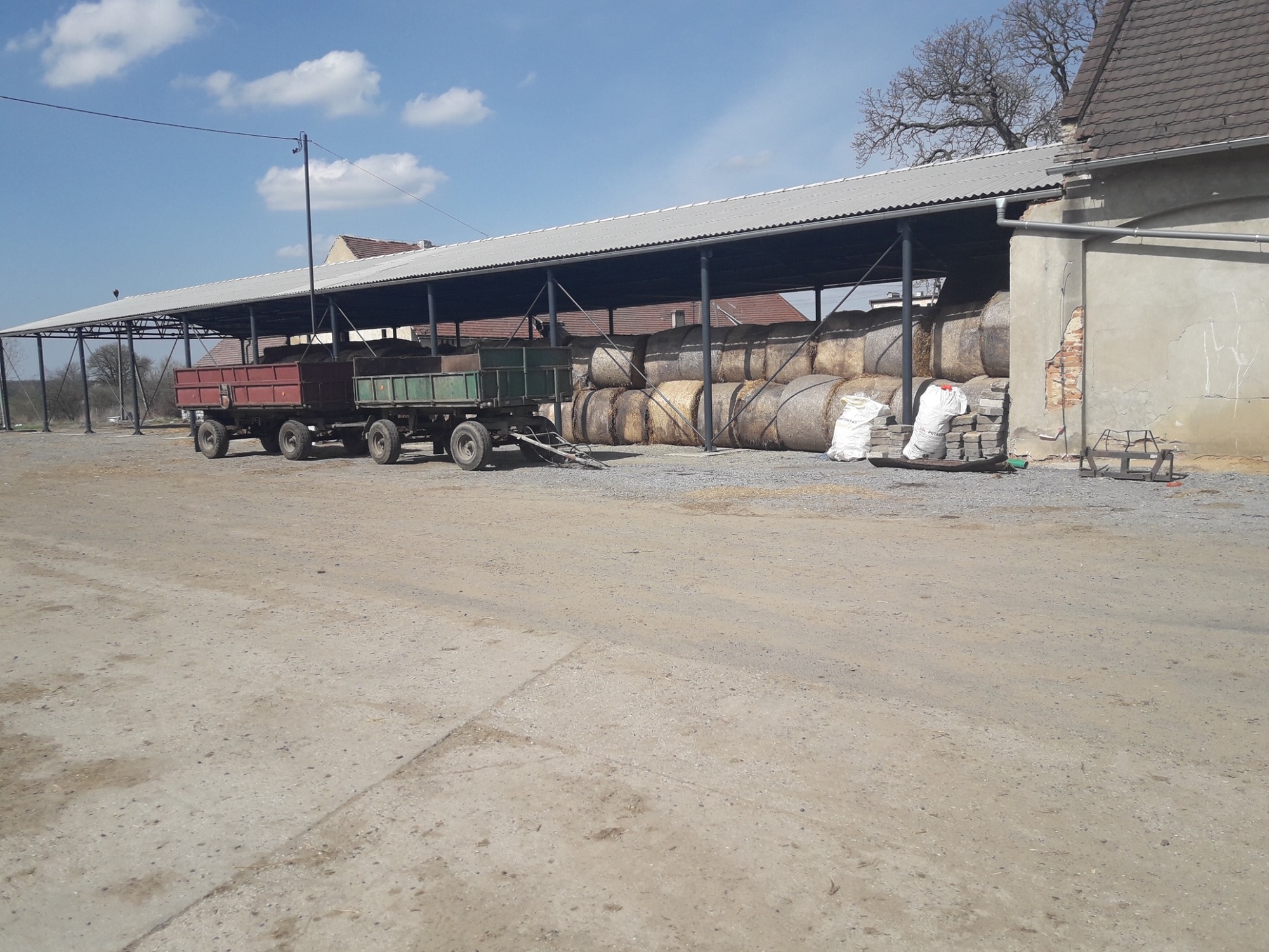 Zadaszona wiata stalowa z wypełnieniem z bel słomy, obok zaparkowana przyczepa rolnicza, teren utwardzony.