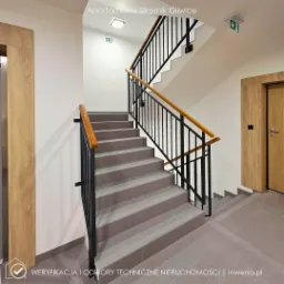 Klatka schodowa w nowoczesnym budynku apartamentowym z szarymi schodami, czarną balustradą i drewnianymi poręczami. Widoczne drzwi i oznaczenia dr&oacute;g ewakuacyjnych.