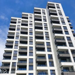 INWERSA Edyta Werkowska - Nowoczesny, biały apartamentowiec z szarymi balkonami i niebieskimi oknami, widziany z dołu pod kątem, na tle błękitnego nieba z delikatnymi chmurami.