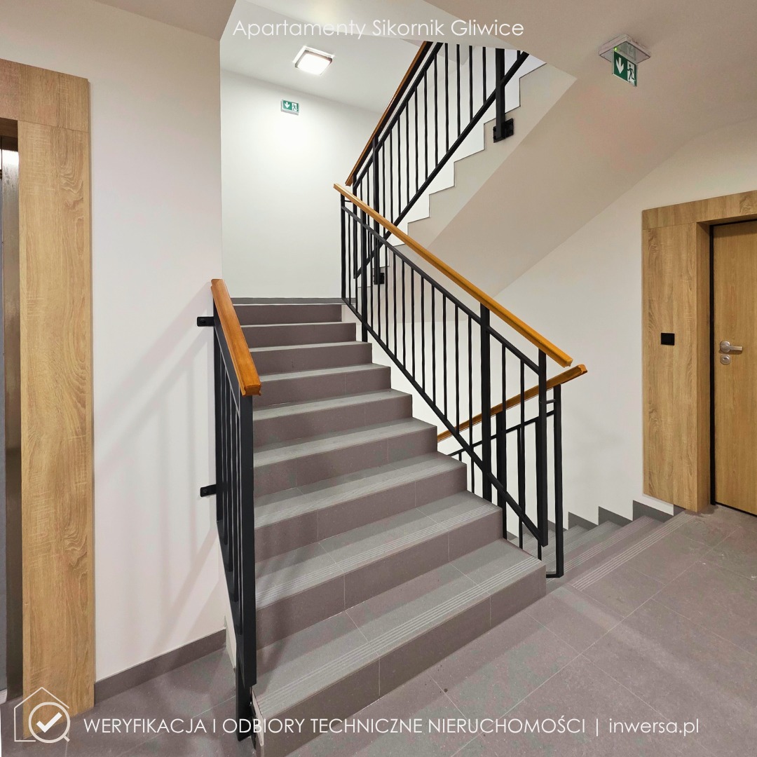 Klatka schodowa w nowoczesnym budynku apartamentowym z szarymi schodami, czarną balustradą i drewnianymi poręczami. Widoczne drzwi i oznaczenia dróg ewakuacyjnych.