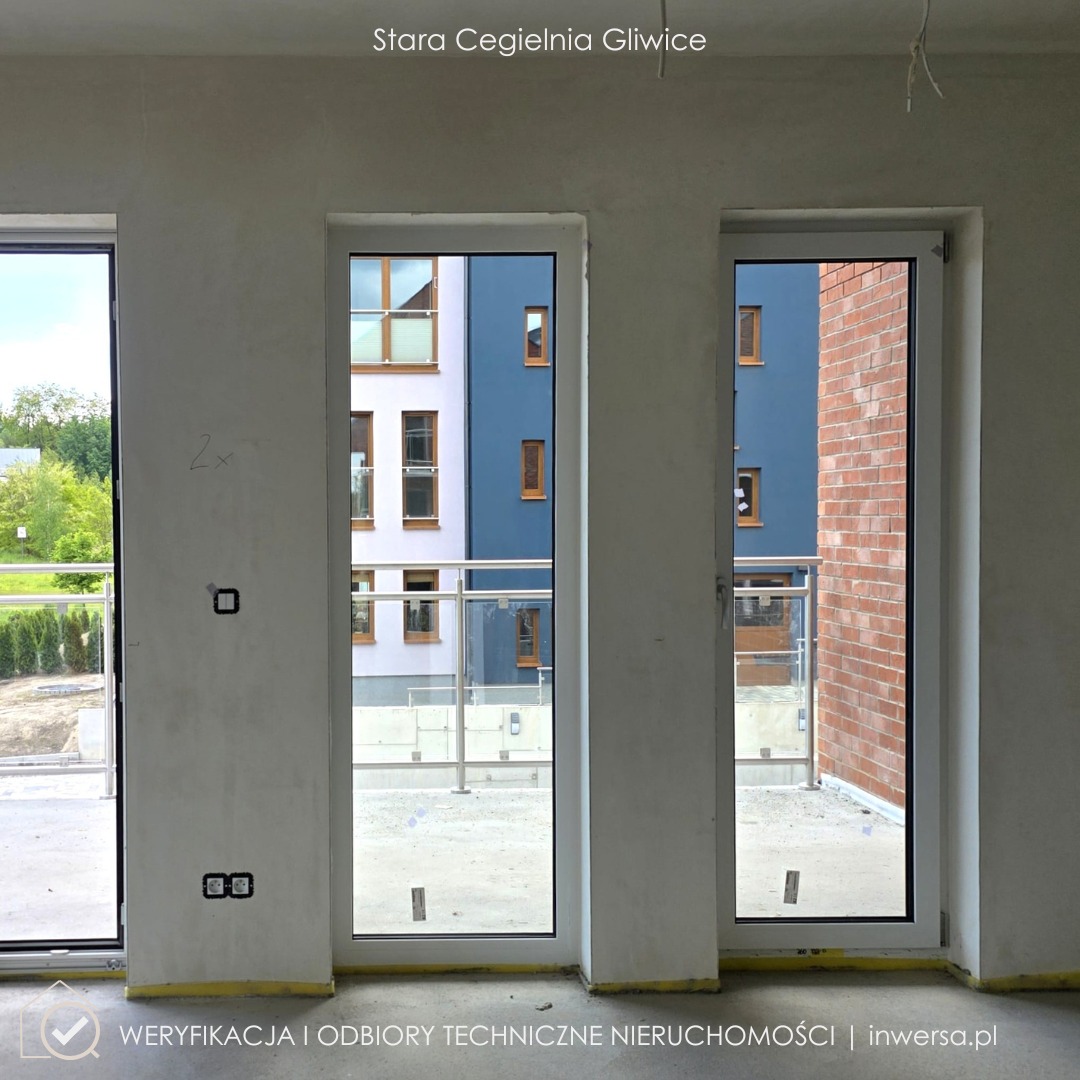 Wnętrze w stanie deweloperskim z trzema oknami, widok na budynki z zewnątrz. Żółta taśma malarska na podłodze, widoczne gniazdka elektryczne. Stara Cegielnia Gliwice.