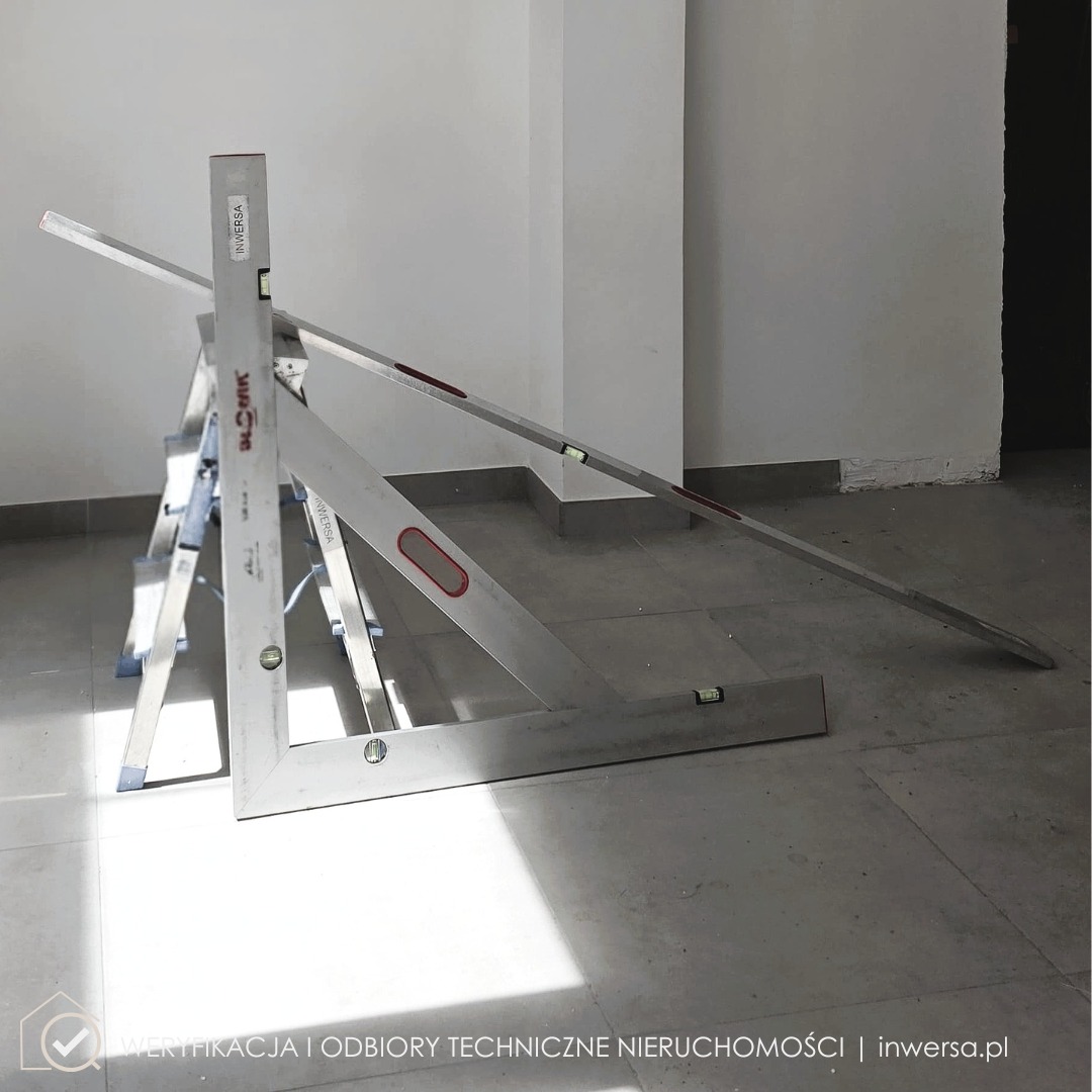 Poziomice i kątownik na schodach w jasnym wnętrzu z szarymi płytkami, przygotowane do pomiarów. Widoczny fragment ściany i wnęka.